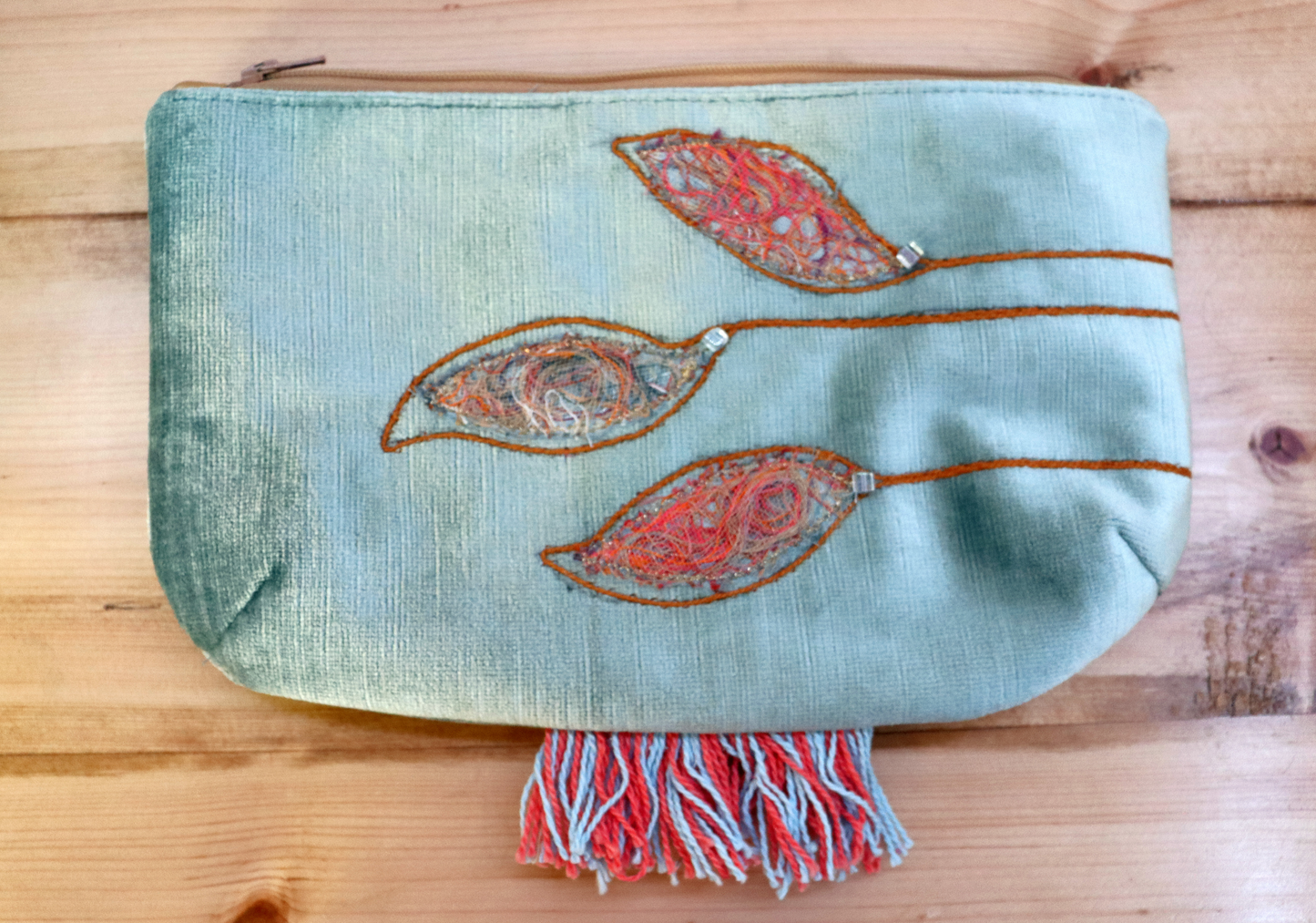 A grayish blue handbag with a floral applique design and decorative stitches, featuring a fringe detail at the bottom.