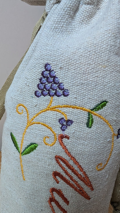 Close up of A cream-colored wine bottle bag with a drawstring closure and a tan drawstring. The bag features a gold embroidered corkscrew and the name "Murray" in gold script.