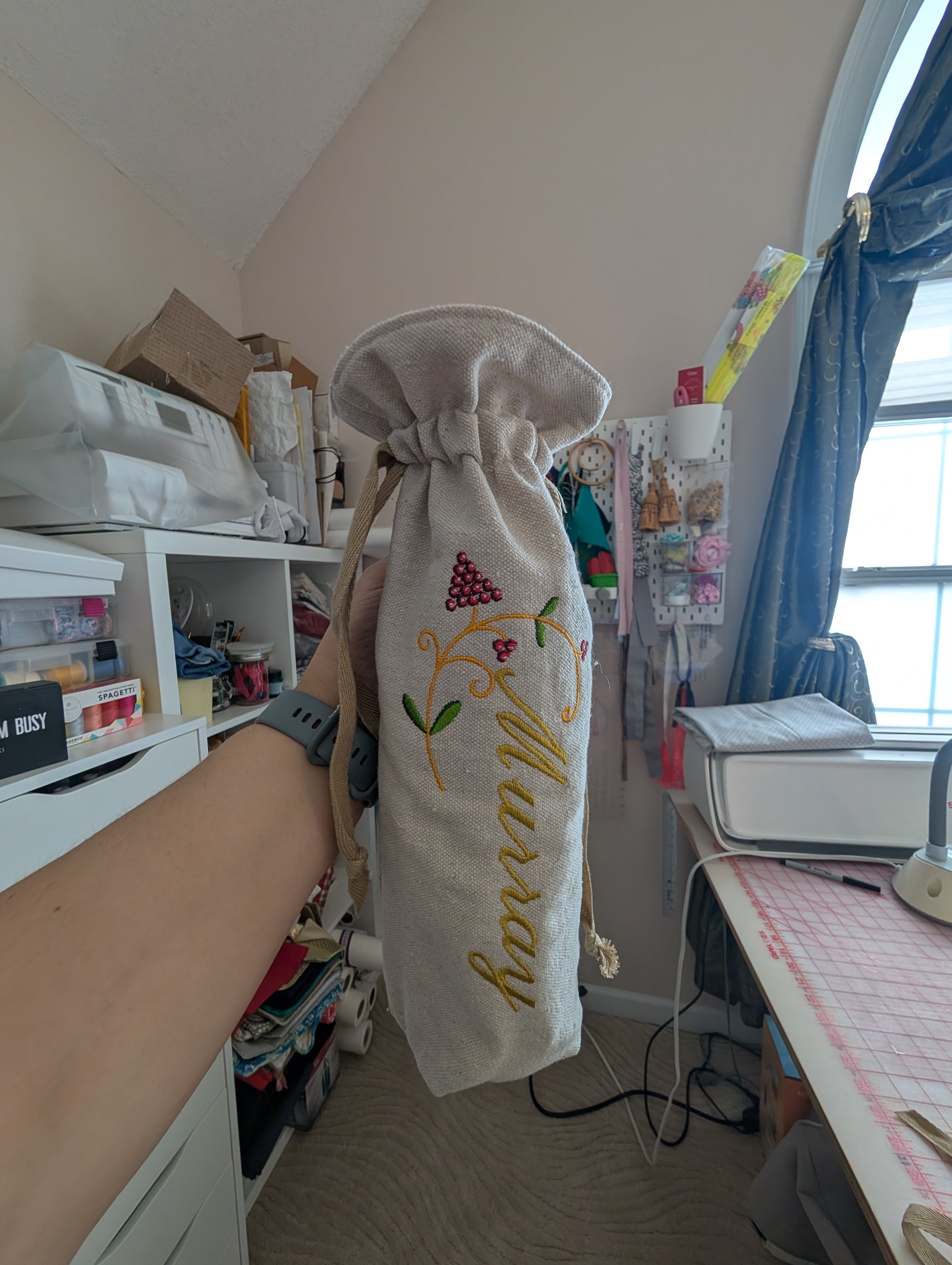 Hand holding a drawstring bag with embroidery in a room with a desk and window.