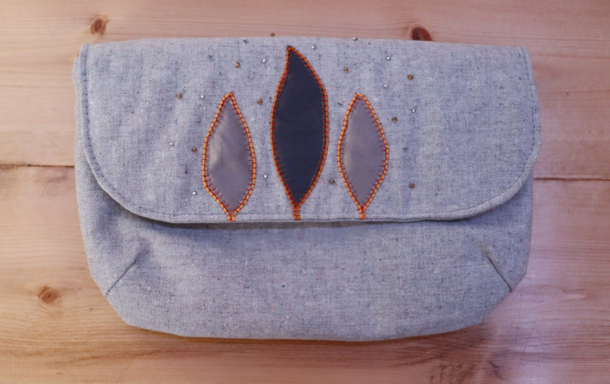A gray handbag with a flap closure, featuring leaf motifs appliquéd in a lighter shade, placed on a wooden surface.